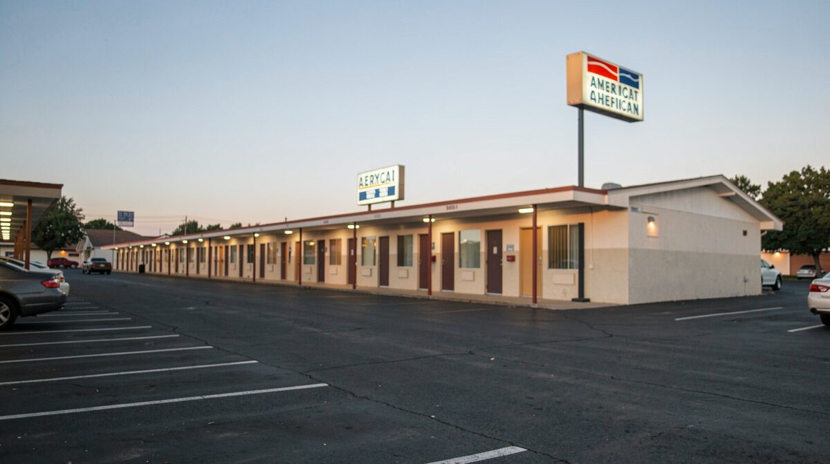 Et langt, lavt amerikansk motel set skråt fra parkeringspladsen i aftenlys, med rækker af ens døre og et oplyst skilt mod en klar himmel